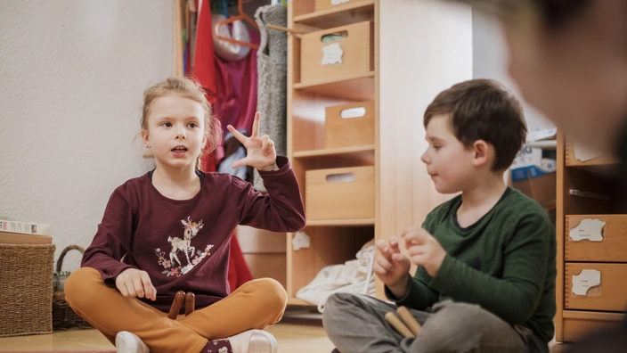 Zwei Kinder sitzen im Kreis auf dem Boden einer Kita und kommunizieren mit Gebärdensprache. Das Bild vermittelt Inklusion, frühkindliche Bildung und spielerisches Lernen in einer wertschätzenden Umgebung.