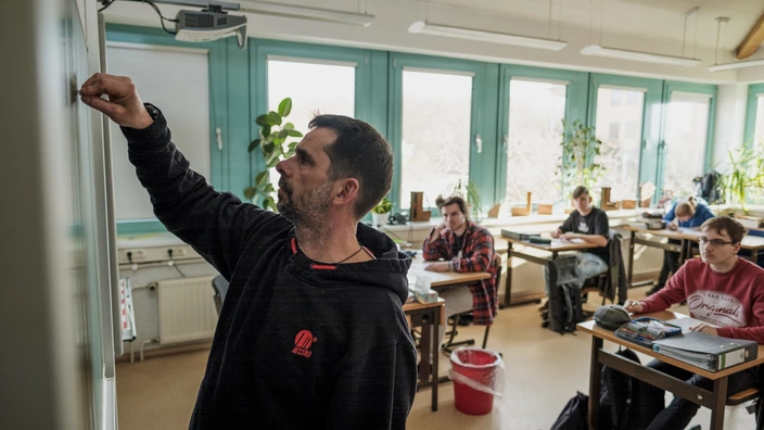 Lehrer schreibt an die Tafel in einem hellen Klassenraum, während mehrere Schülerinnen und Schüler aufmerksam an ihren Tischen sitzen und arbeiten. Das Bild vermittelt modernen Unterricht, Lernatmosphäre und individuelle Förderung in der Schule.