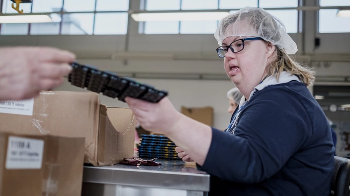 Frau mit Down-Syndrom arbeitet konzentriert an einem Packtisch in einer Werkstatt, trägt Haarnetz und Brille. Das Bild zeigt inklusive Beschäftigung, Teilhabe am Arbeitsleben und selbstständiges Arbeiten in der Produktion.